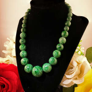 Angled view of a graduated green Maw Sit Sit jade bead necklace on a black jewelry bust with floral accents. The lighting highlights the stone’s deep green cosmic hue and smooth glassy surface
